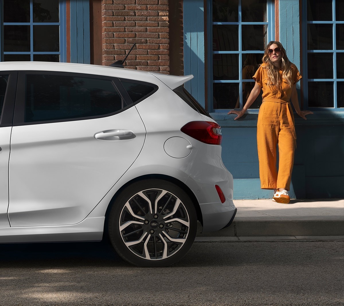 Ford Fiesta in Weiss. Seitenansicht des Hecks, vor einem Haus parkend mit Frau im Hintergrund.