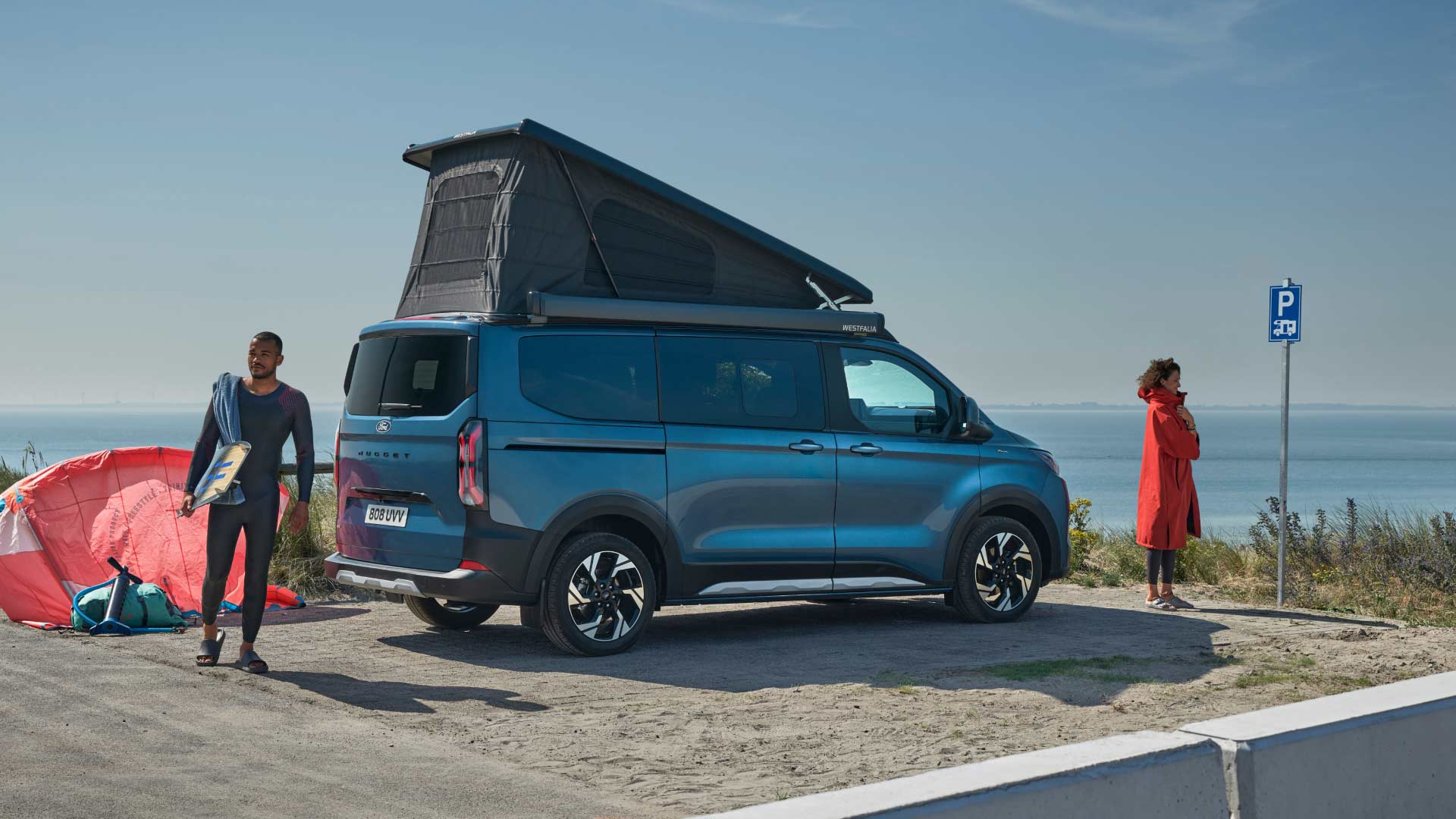 Ein blauer Ford Transit Custom Camper Van mit Aufstelldach ist am Meer geparkt, mit zwei Personen und einem Surfbrett in der Nähe. 