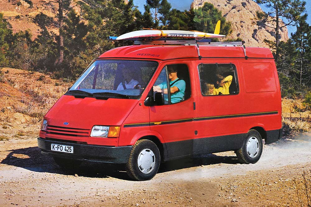 Ford Nugget von 1993 mit Dachträger und Surfbrett fährt auf staubiger Bergstrasse durch die Berge.