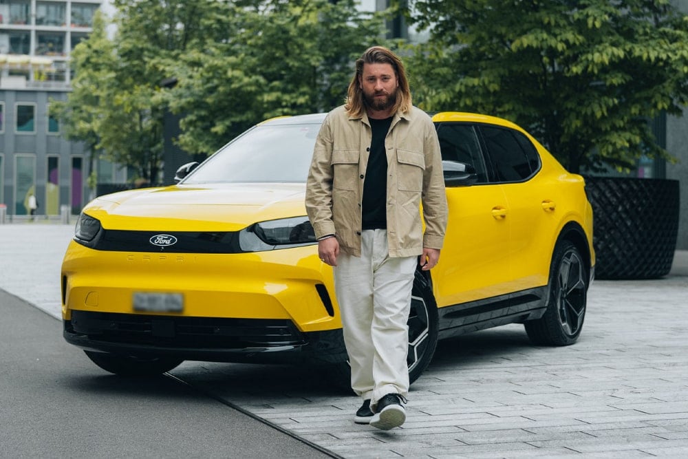 Mann mit langen Haaren läuft cool vor einem gelben Ford Capri