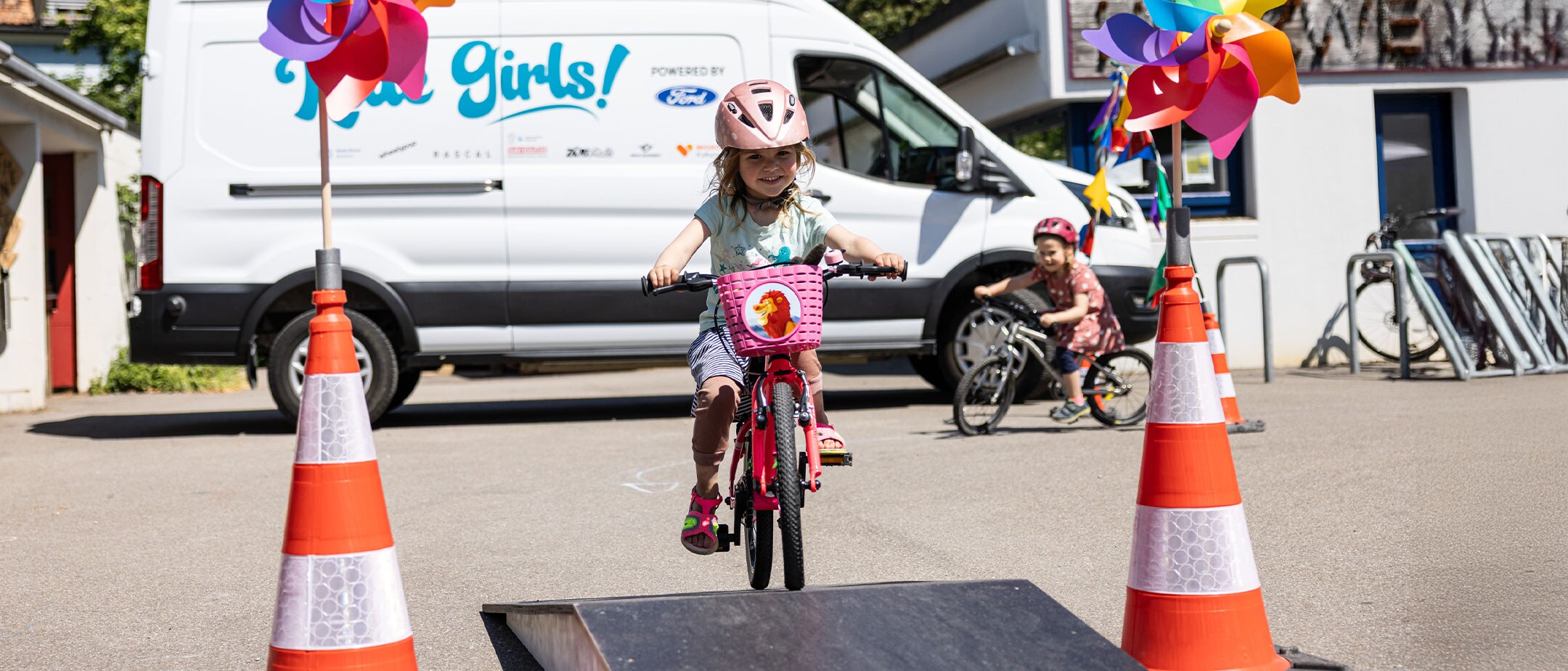 Ein kleines Mädchen fährt mit einem Fahrrad auf einem Hindernis-Parcours. Im Hintergrund steht ein Ford E-Transit