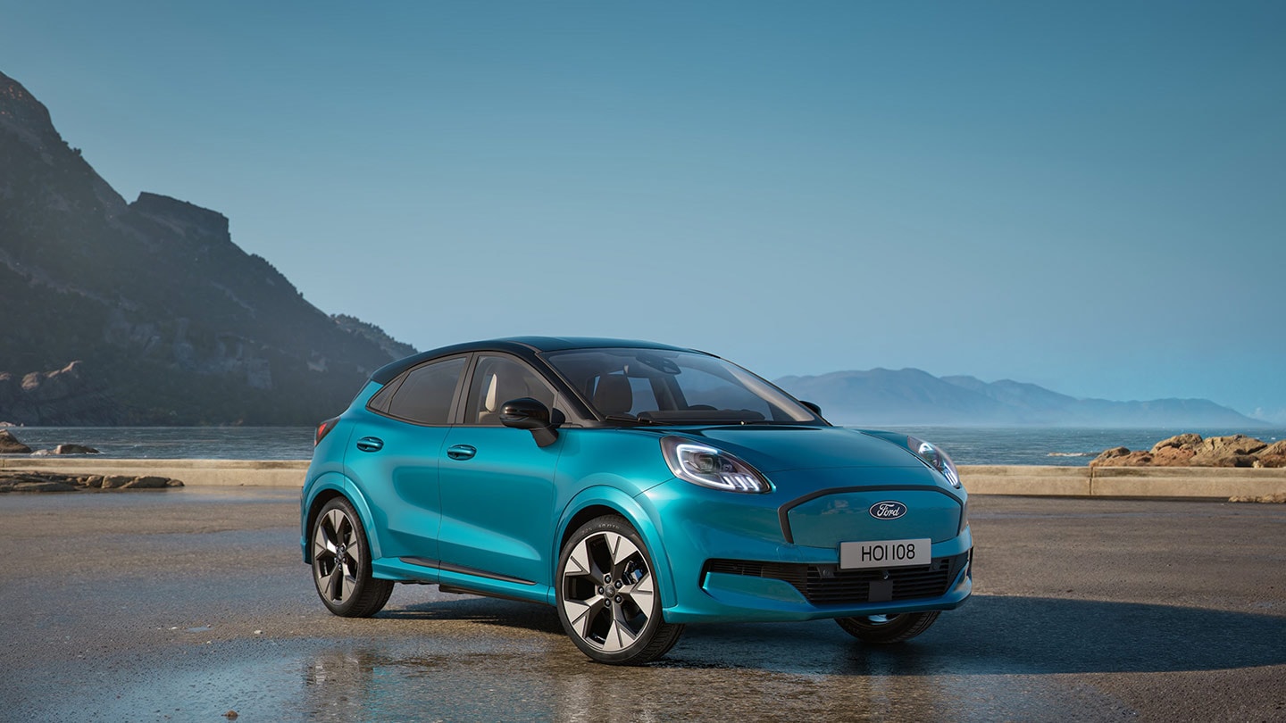 Vorderansicht eines blauen Ford Puma Gen-E, parkiert in der Nähe eines Gewässers, mit Bergen im Hintergrund.