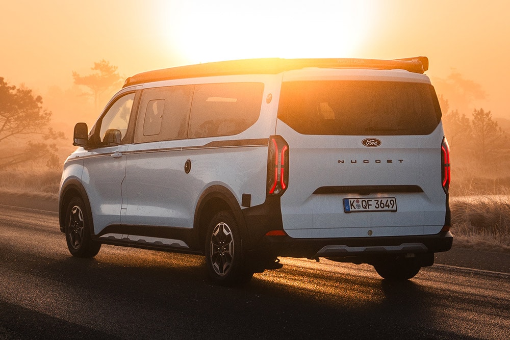 Heckansicht der Ford Nugget Jubiläumsedition auf einer asphaltierten Strasse in einer nebligen Landschaft bei Sonnenaufgang.