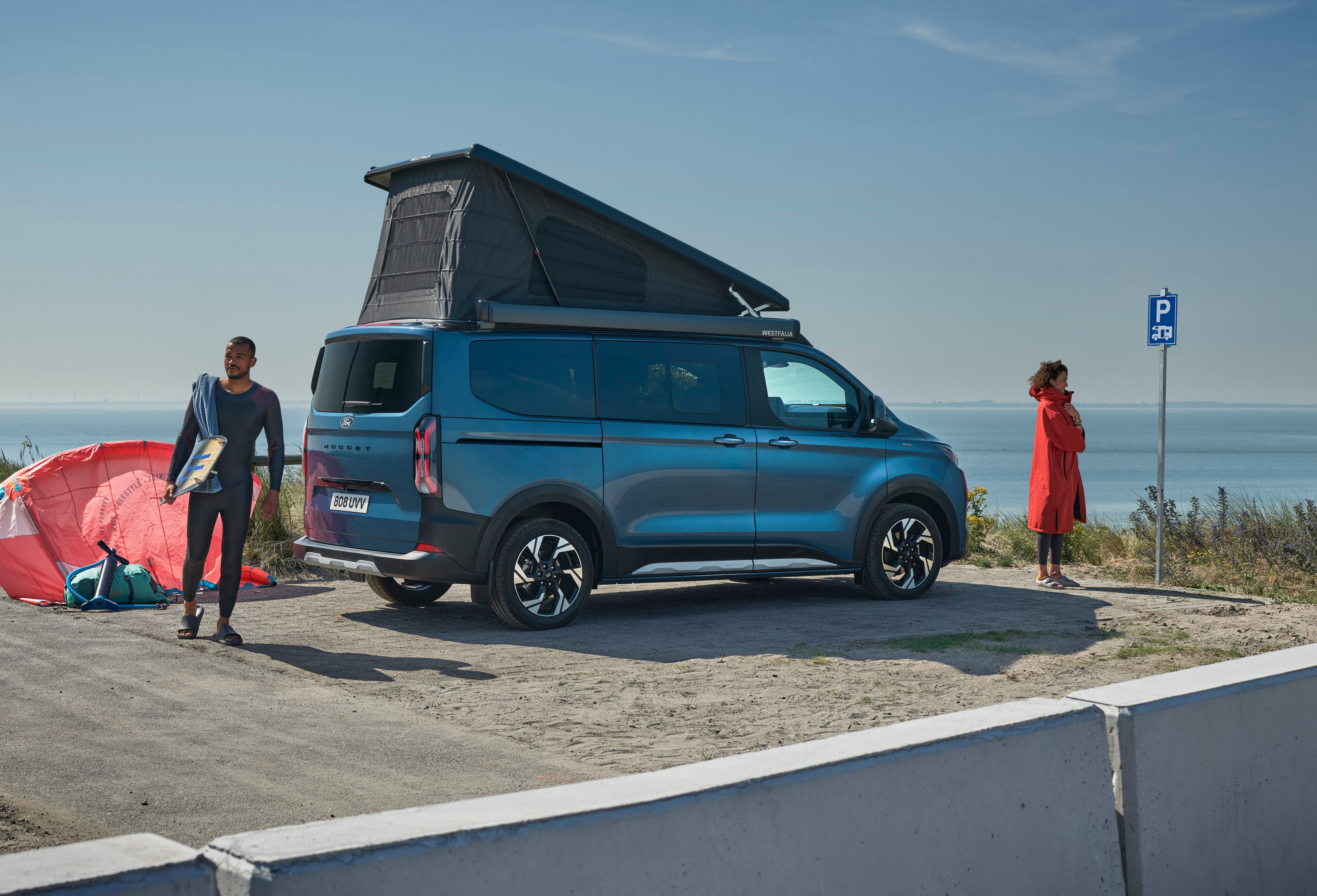 Ein dunkelblauer Ford Nugget mit aufgestelltem Dach ist am Meer parkiert, während zwei Personen Kitesurf-Ausrüstung bereitlegen.