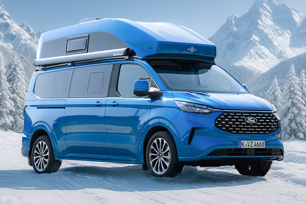 Ein blauer Ford Nugget mit Hochdach steht im Schnee vor einer Bergkulisse, aus der vorderen Dreiviertelansicht.
