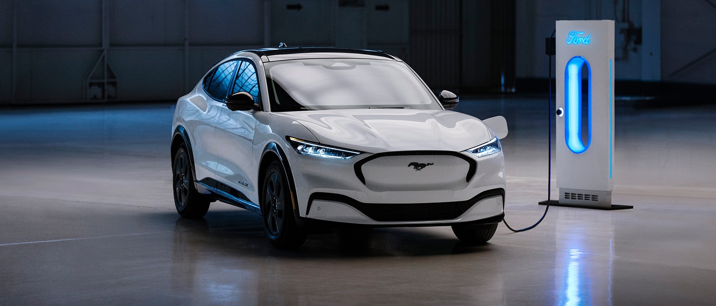 Ein weisser Ford Mustang Mach-E steht in einer Garage und ist an eine blau leuchtende Ladestation angeschlossen..