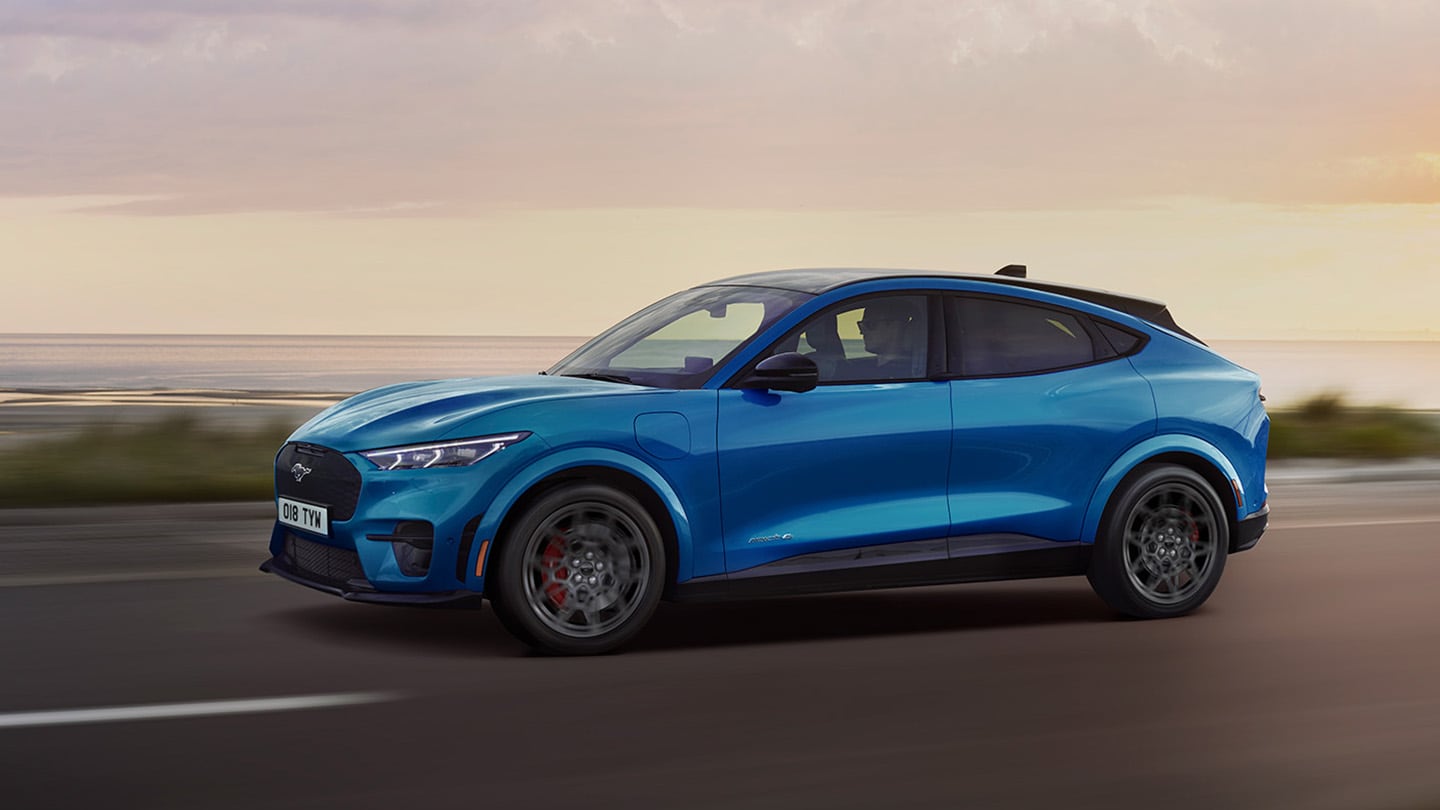 Seitenansicht eines blauen elektrischen Ford Mustang Mach-E, der in der Abenddämmerung eine Küstenstrasse entlangfährt.