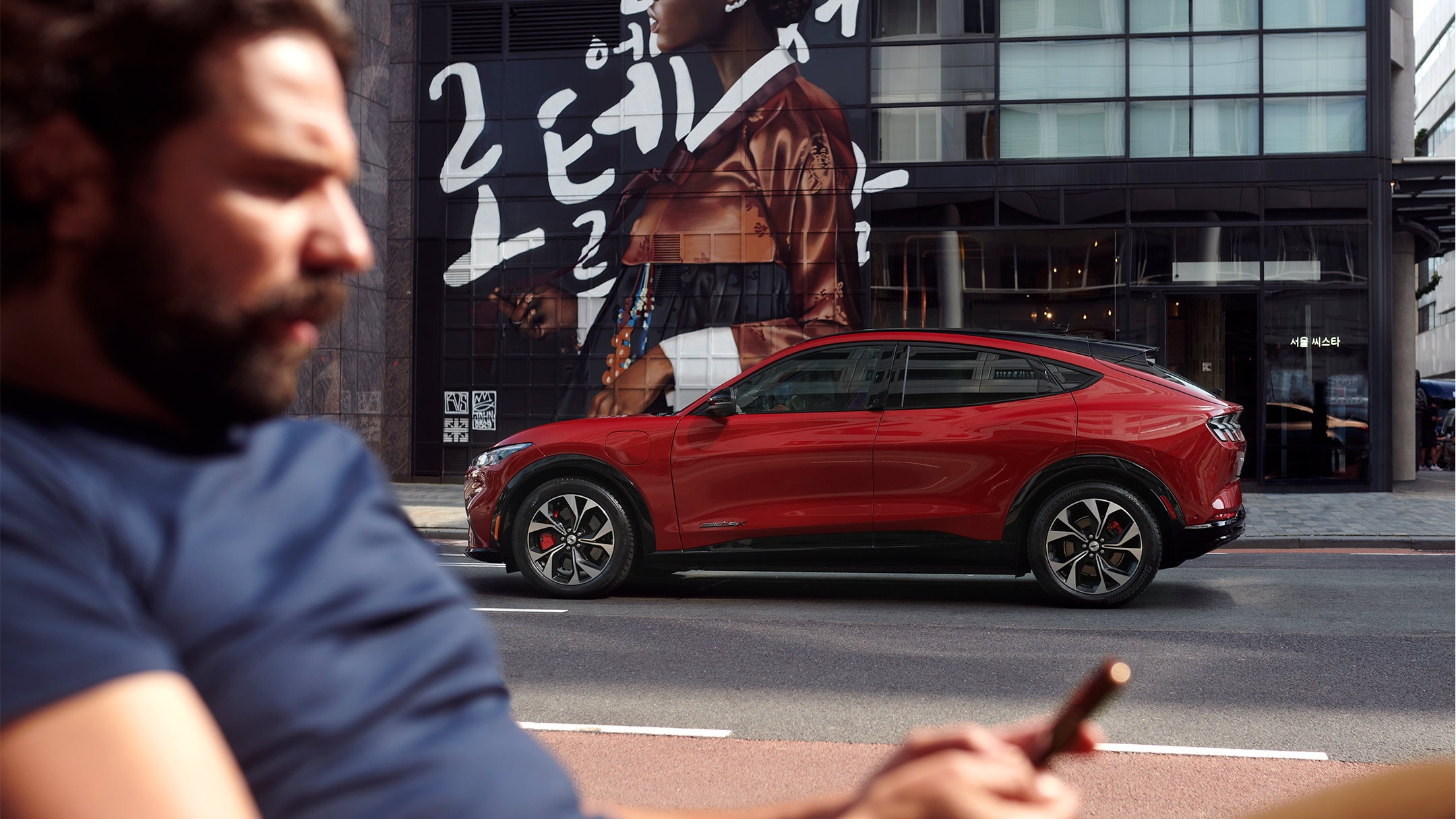 Ford Mustang Mach-E in Rot. Seitenansicht. Im Vordergrund ein Mann der auf sein Smartphone schaut