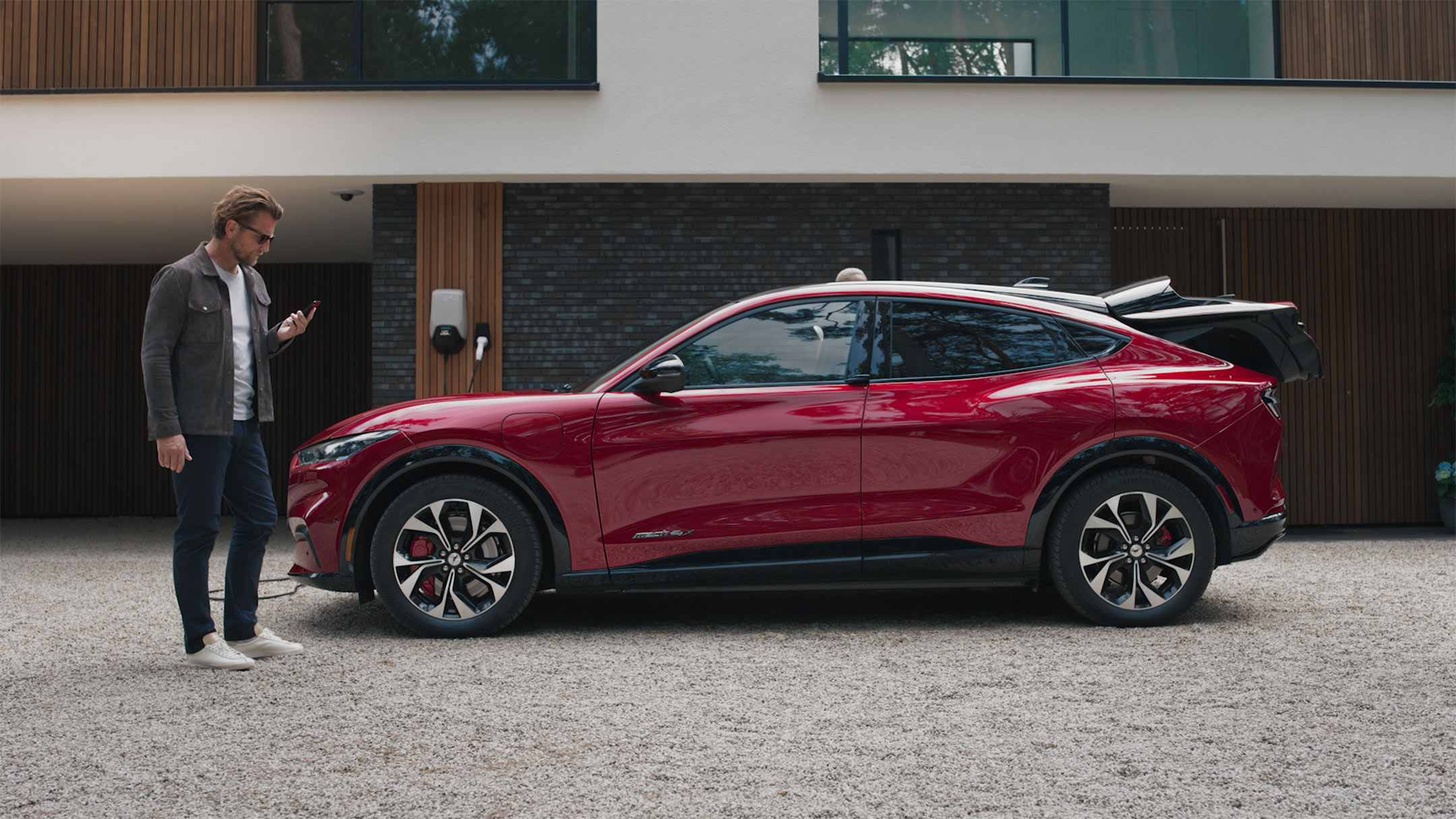 Ford Mustang Mach-E in Rot. Seitenansicht. Im Vordergrund ein Mann der auf sein Smartphone schaut