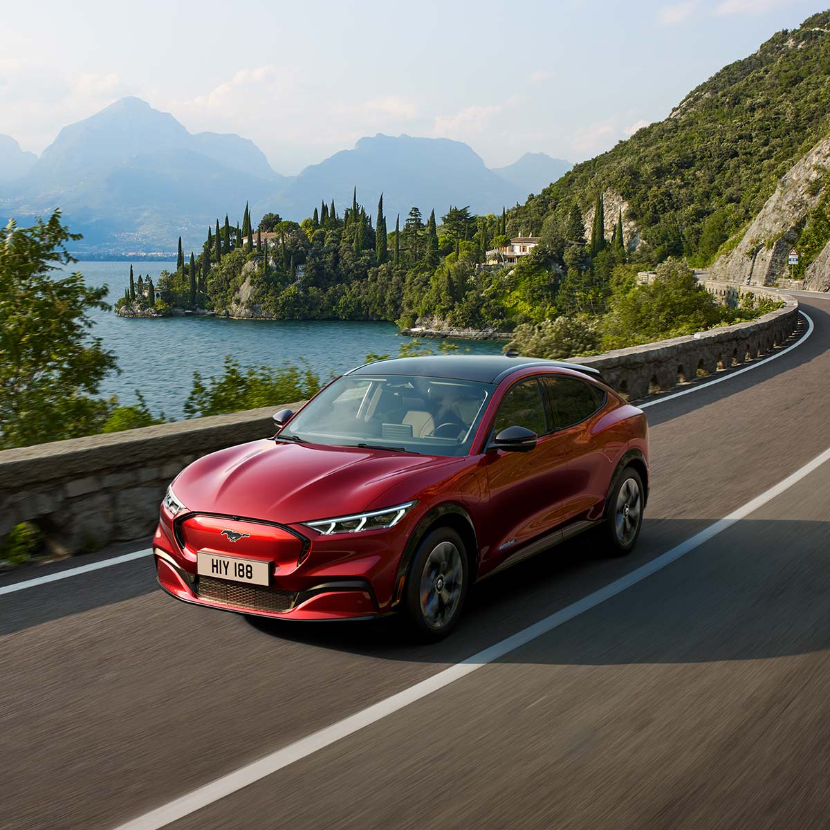 Ford Mustang Mach-E in Rot. Dreiviertelansicht, fahrend auf Bergstrasse vor einem See