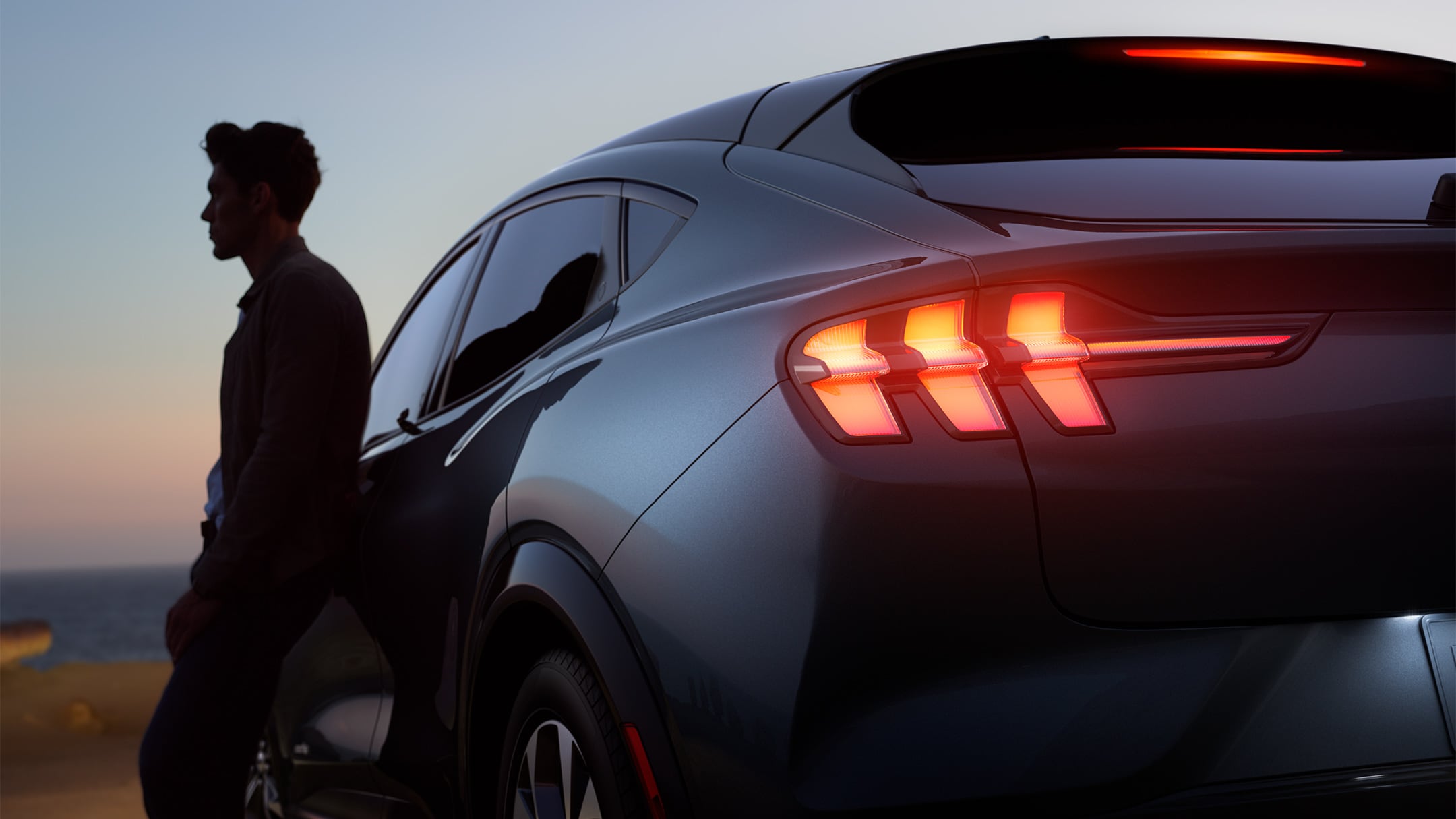 Ford Mustang Mach-E in Grau. Mann lehnt in Abenddämmerung am Fahrzeug