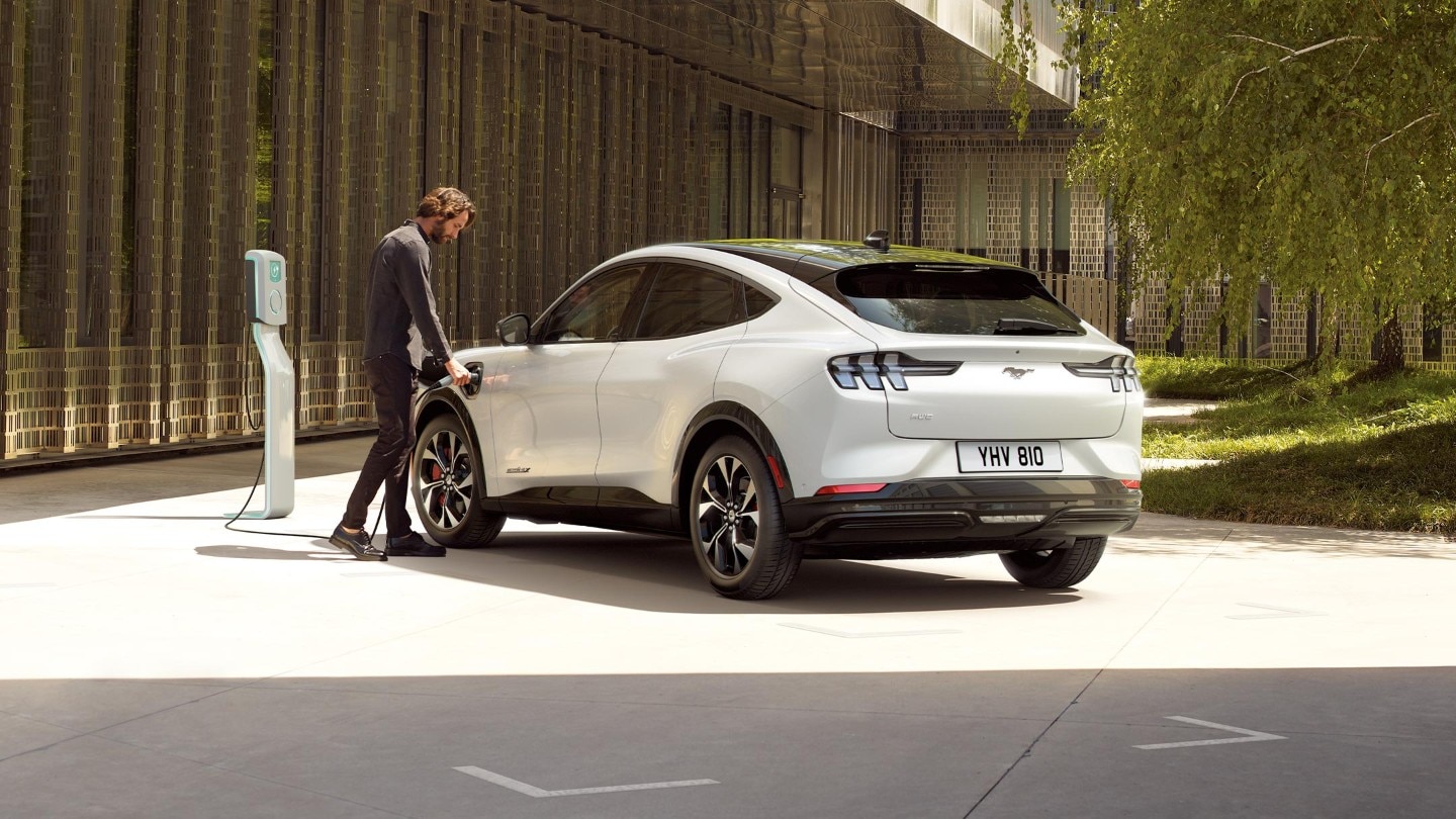 Ford Mustang Mach-E in Weiss. 3/4-Heckansicht. Mann verbindet die Ford Connected Wallbox mit dem Ladeanschluss des Fahrzeugs