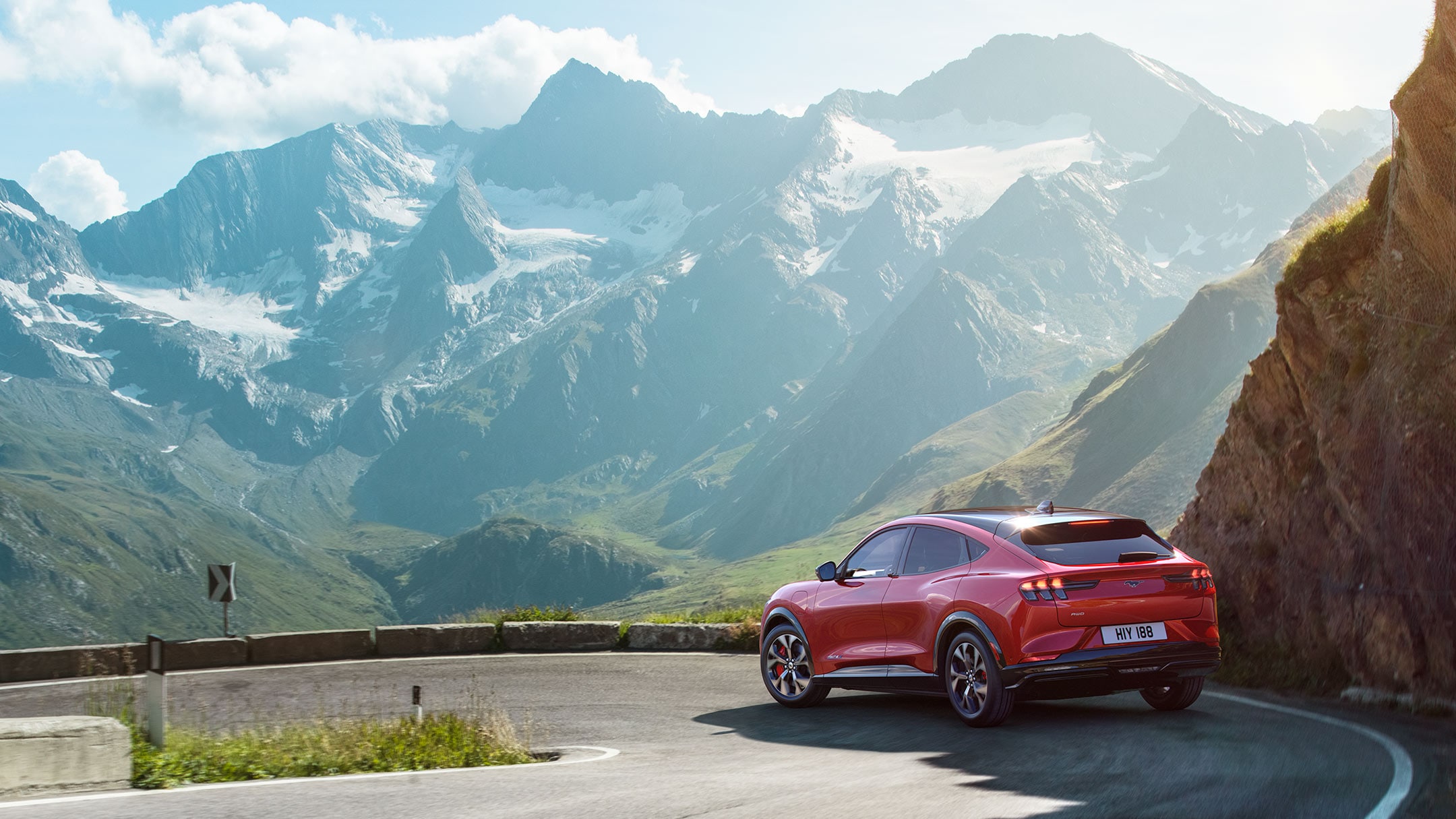 Ford Mustang Mach-E in Rot. 3/4-Heckansicht. Fahrszene auf einer kurvigen Bergpanorama-Strasse