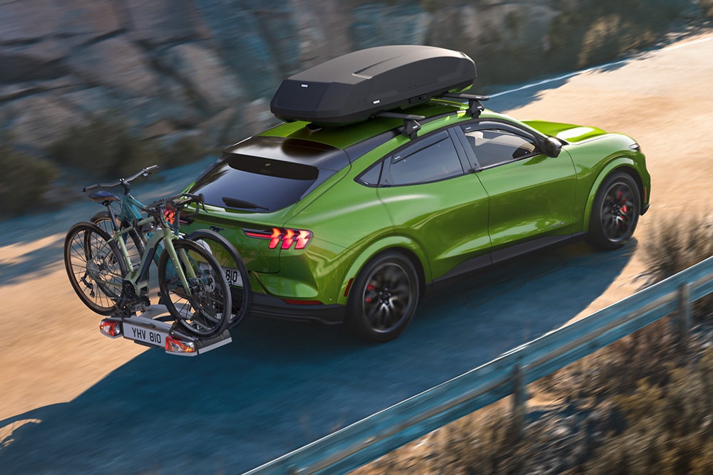 Ford Mustang Mach-E in Grün mit Fahrradträger und Dachbox auf einer kurvenreichen Straße.