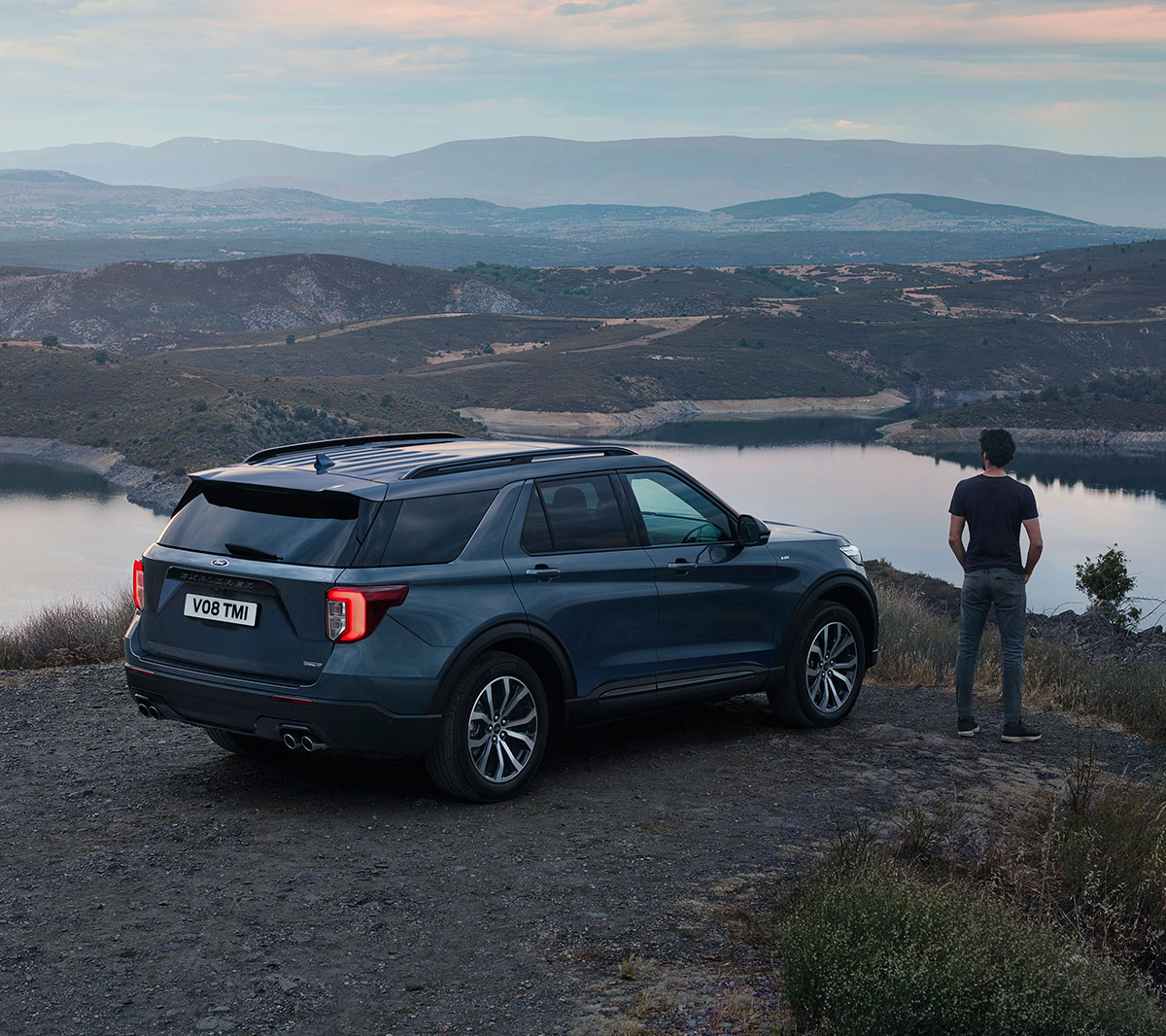 Ford Explorer Plug-In Hybrid in Blau. Dreiviertel-Heckansicht. In hügeliger Landschaft parkend mit nebenstehender Person die Landschaft betrachtend