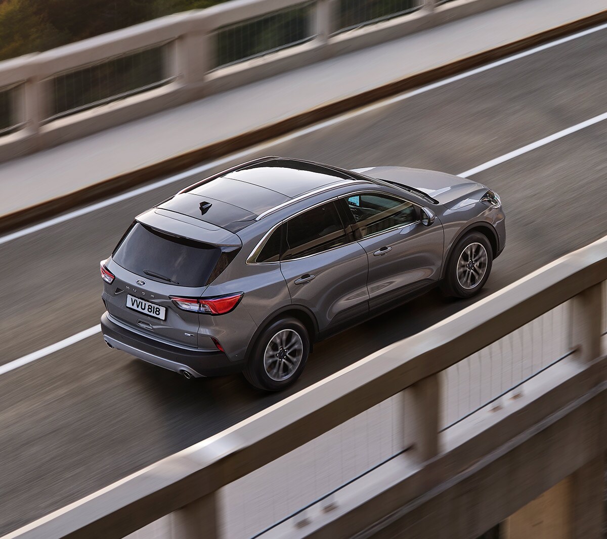 Ford Kuga in Grau. 3/4-Heckansicht von oben, über eine Brücke mit Wald im Hintergrund fahrend