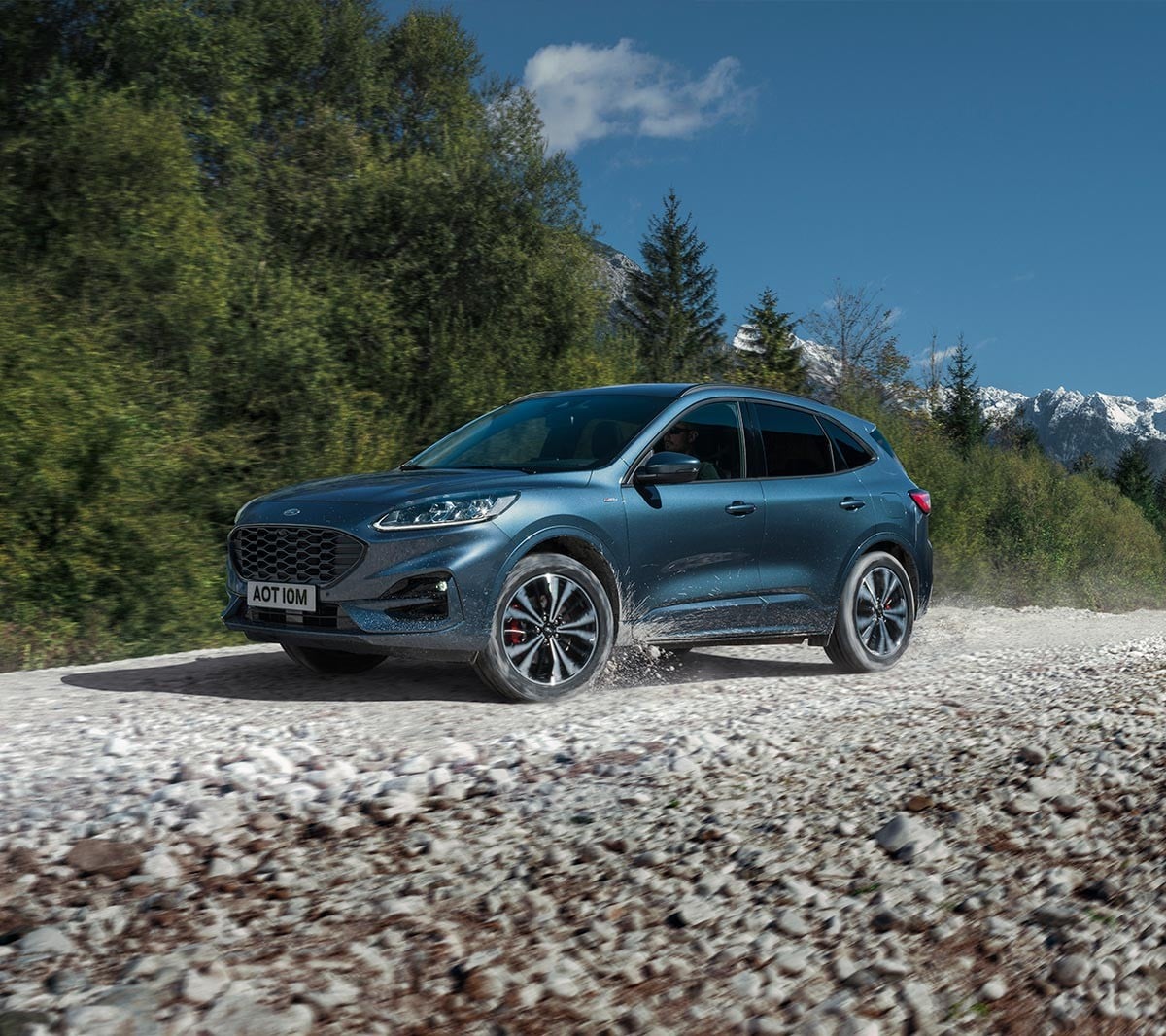 Ford Kuga in Blau. 3/4-Frontansicht auf Schotterweg vor einem Wald fahrend