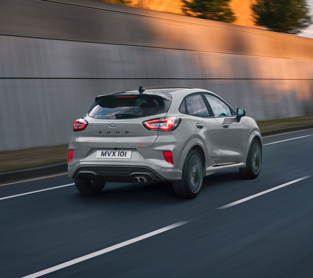 Ford Puma ST in Grau. ¾-Frontansicht auf einer Strasse vor modernem Gebäude. Eine Frau im Hintergrund