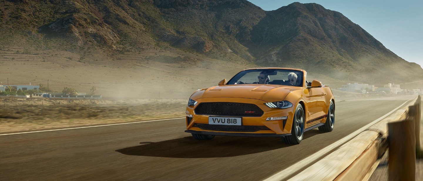 Ford Mustang California in Orange. Dreiviertel-Frontansicht, in der Natur vor Bergen im Hintergrund fahrend.