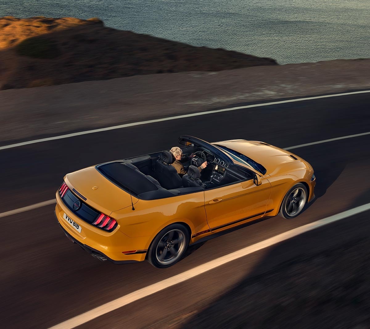 Ford Mustang California in Orange. Dreiviertel-Heckansicht, auf einer Strasse fahrend.