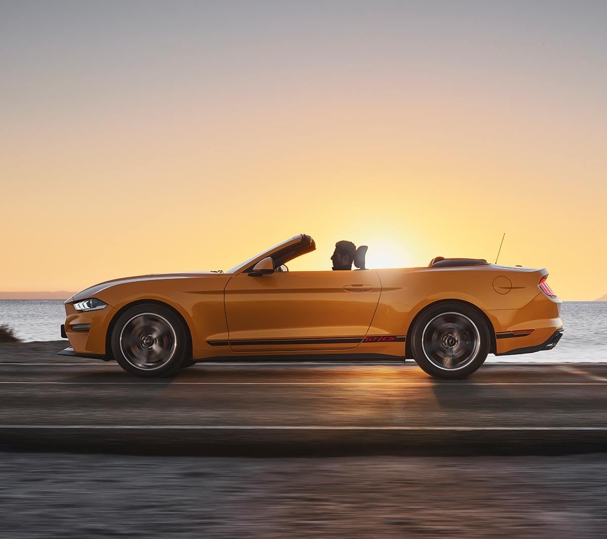 Ford Mustang California in Orange. Seitenansicht, in der Natur im Sonnenuntergang fahrend.