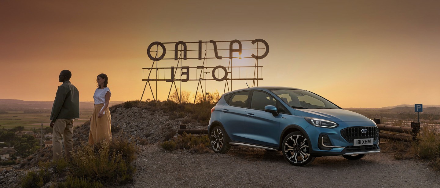 Ford Fiesta in Blau. Dreiviertel-Frontansicht in der Natur parkend. Zwei Personen stehen daneben.