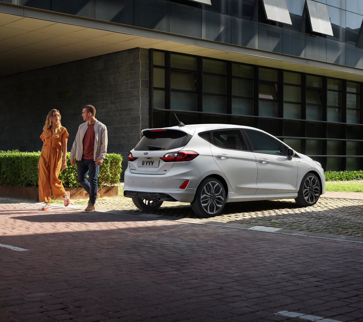 Ford Fiesta in Weiss. Dreiviertel-Heckansicht vor Gebäuden parkend mit zwei Personen daneben.