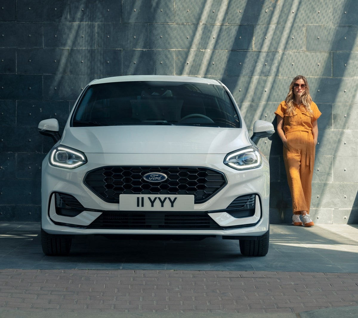 Ford Fiesta in Weiss. Frontansicht vor einem modernen Gebäude parkend. Eine Frau steht daneben.