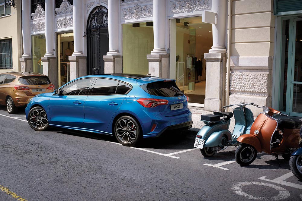 Ein blauer Ford Focus parkiert mithilfe des aktiven Parkassistenten in eine Parklücke in der Stadt.