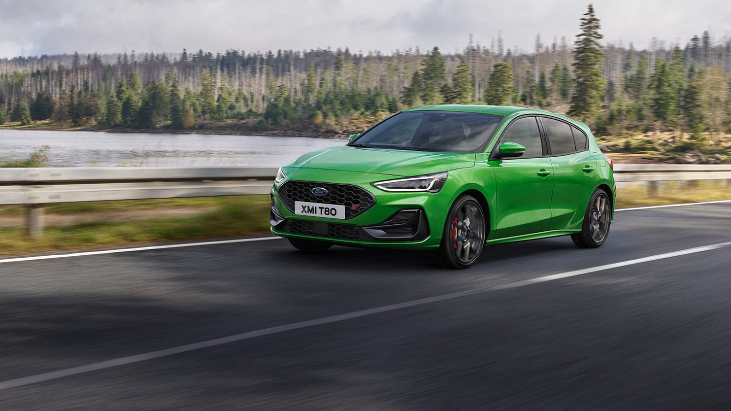 Ein Ford Focus ST in der Farbe «Mean Green» fährt auf einer Strasse, mit Bäumen im Hintergrund.