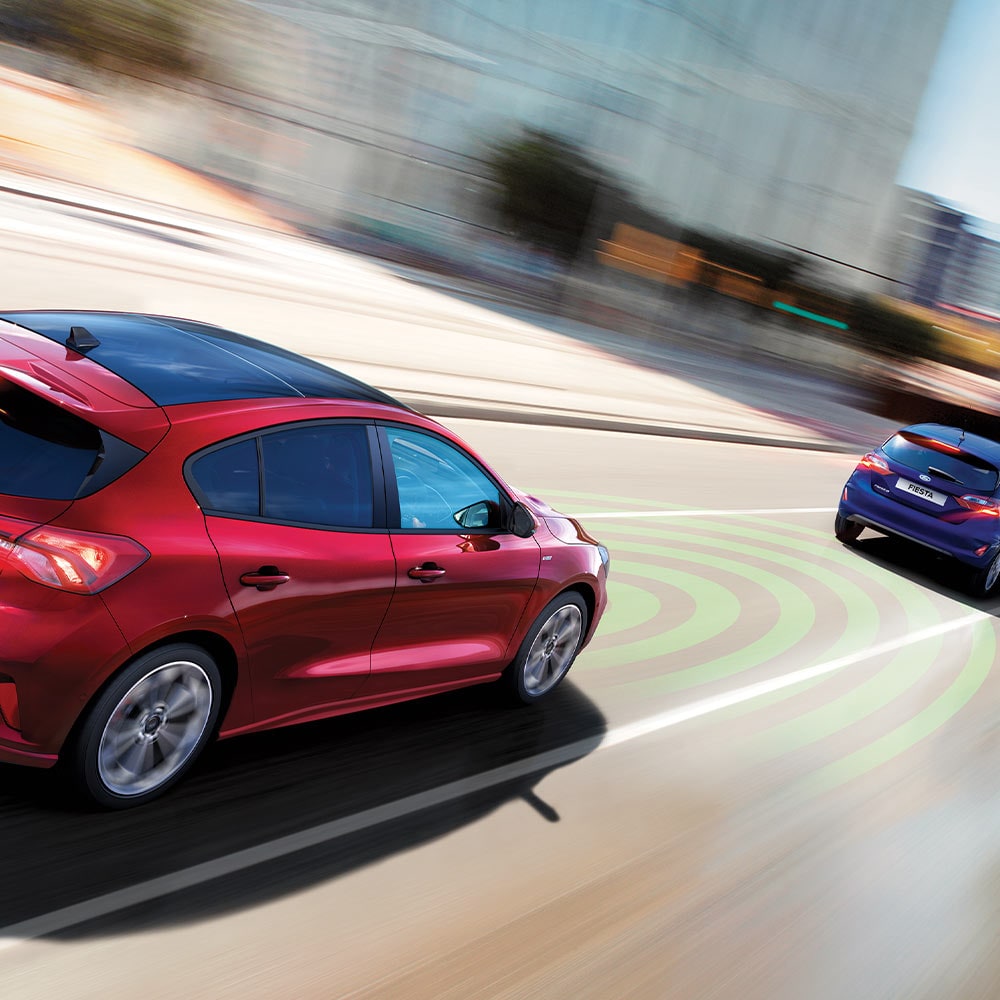 Eine Grafik zeigt einen roten Ford Focus, dessen Sensoren (dargestellt durch blaue Linien) das vordere Fahrzeug erkennen, um das Notbremssystem zu aktivieren.