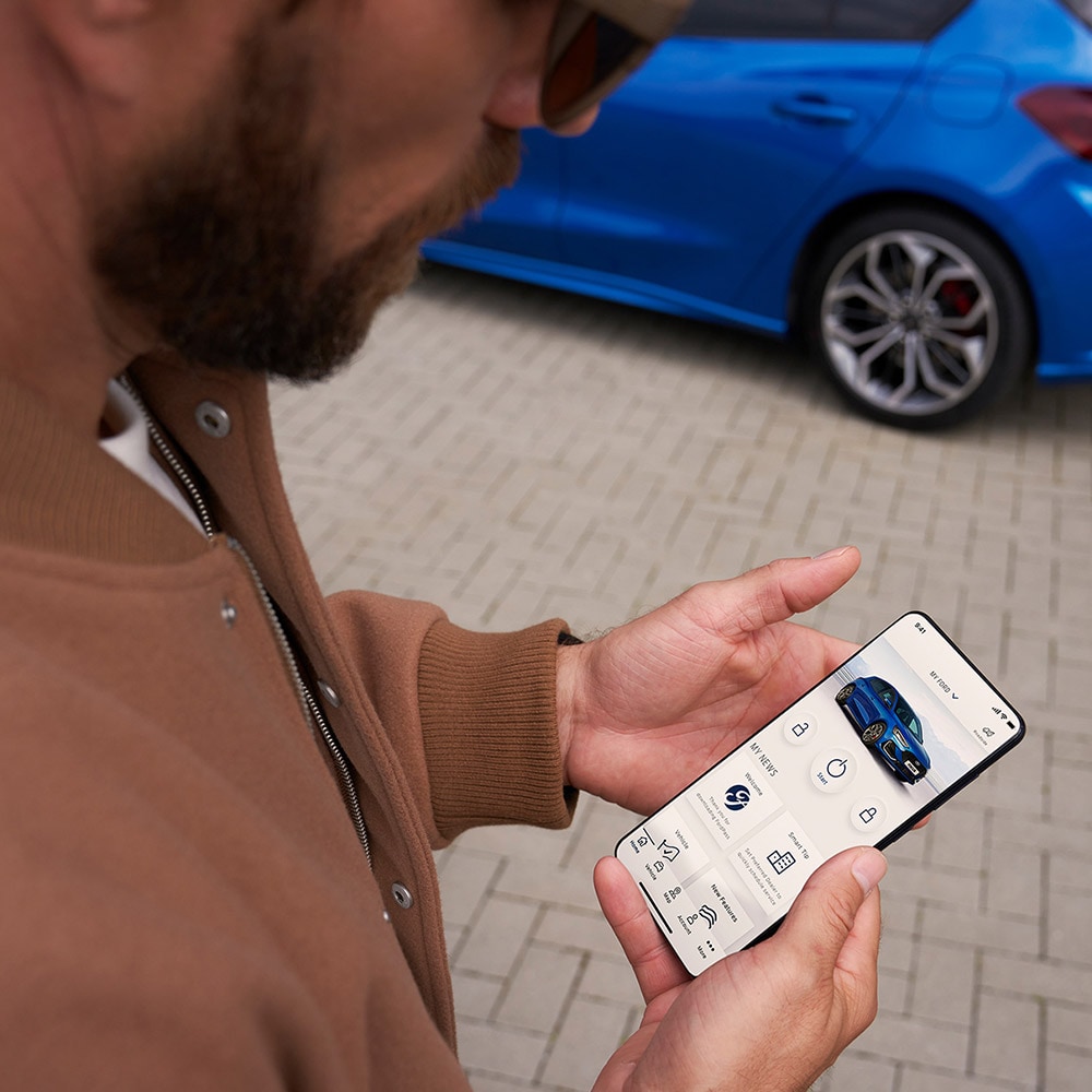 Eine Person hält ein Smartphone, auf dem die FordPass™-App angezeigt wird. Im Hintergrund befindet sich ein blauer Ford Focus.