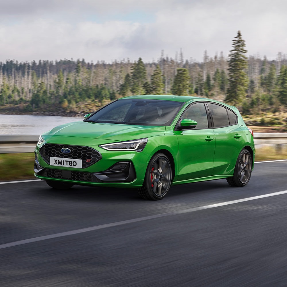 Ein Ford Focus ST in Mean Green fährt auf einer kurvenreichen Strasse umgeben von Bäumen.