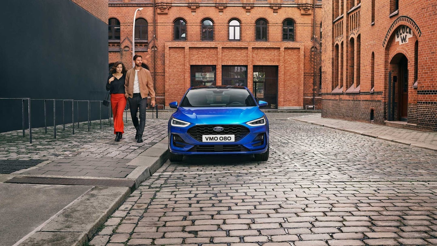 Ford Focus in Blau. Frontansicht, in einer industriellen Gegend parkend. Zwei Personen gehen daneben.