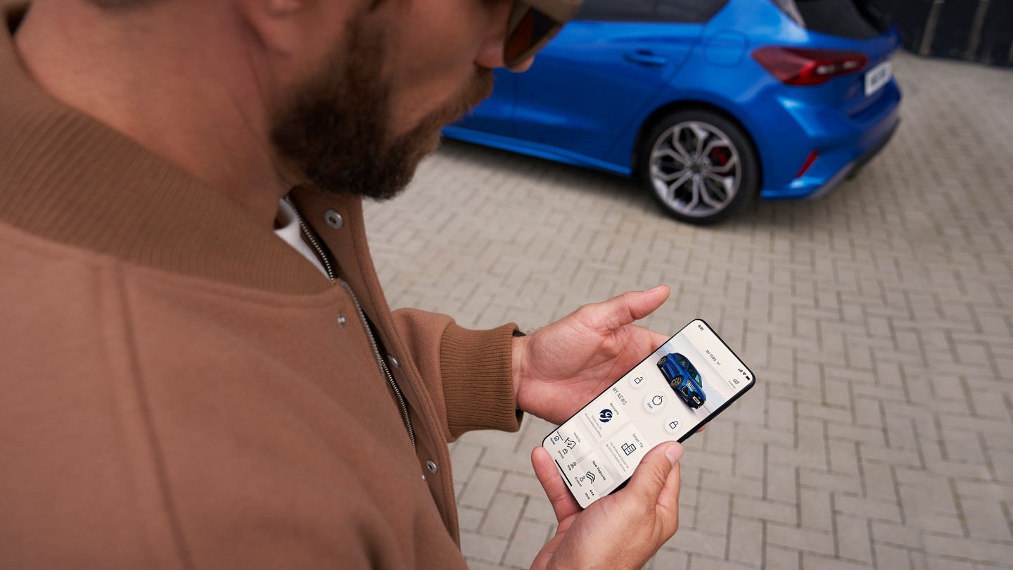 FordPass App. Ein Mann schaut auf sein Smartphone. Im Hintergrund ein blauer Ford Focus.