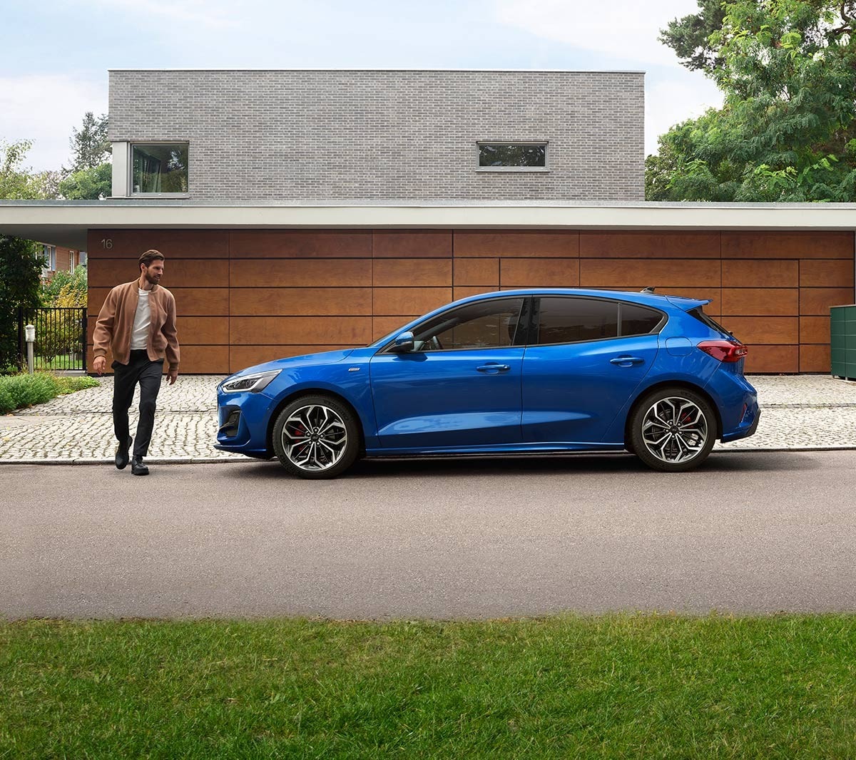 Ford Focus in Blau. Seitenansicht, vor einem modernen Haus parkend. Ein Mann läuft daneben.