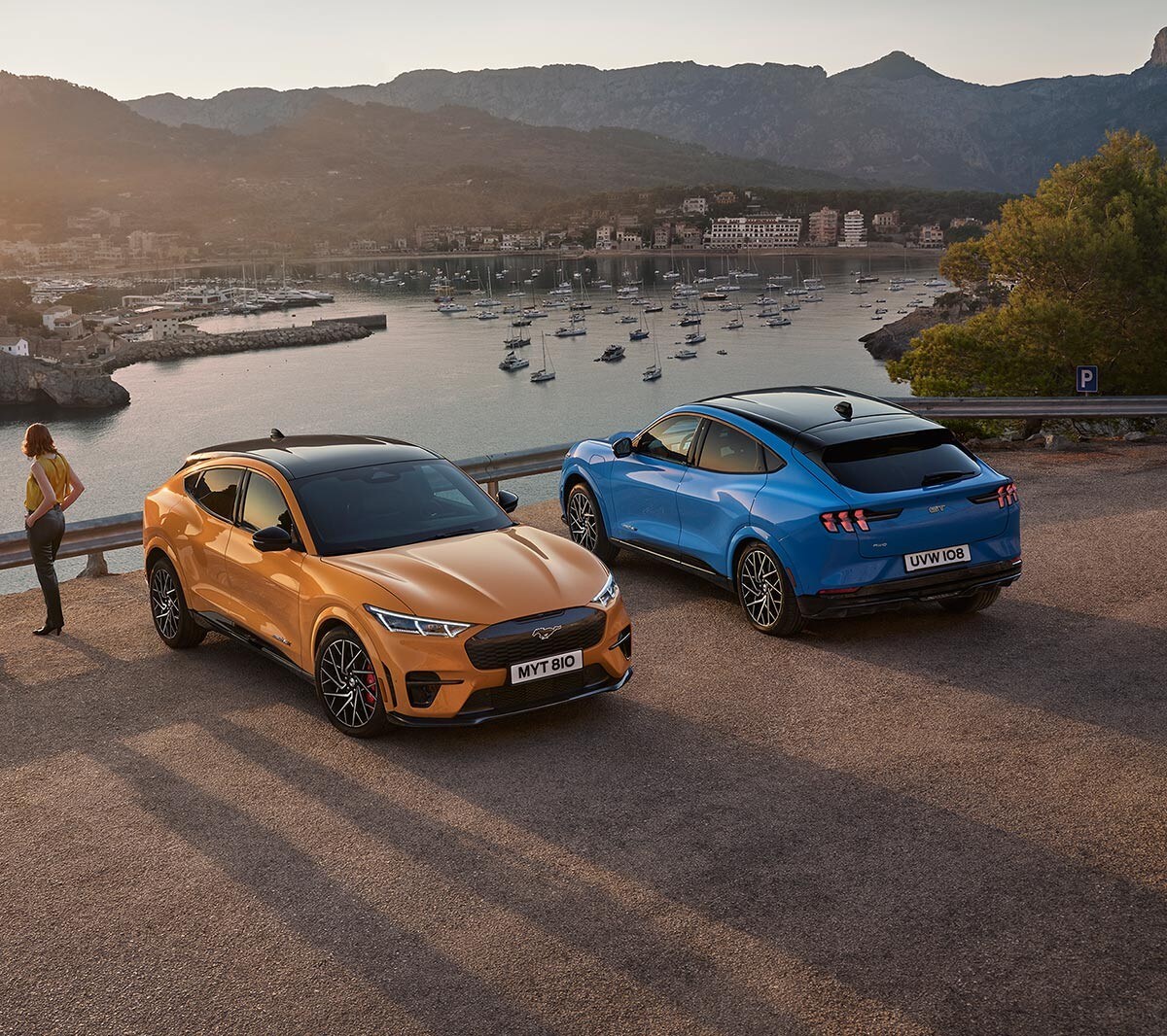 Ford Mustang Mach-E GT in Orange neben einem Ford Mustang Mach-E GT in Blau parkend. Im Hintergrund ein See und Berge im Sonnenuntergang.