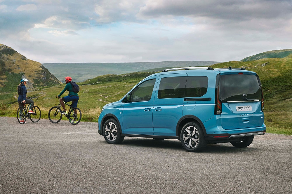 Ein blauer Ford Tourneo Connect ist auf einer Strasse in einer grünen, hügeligen Landschaft parkiert, mit zwei Personen auf Fahrrädern in der Nähe.