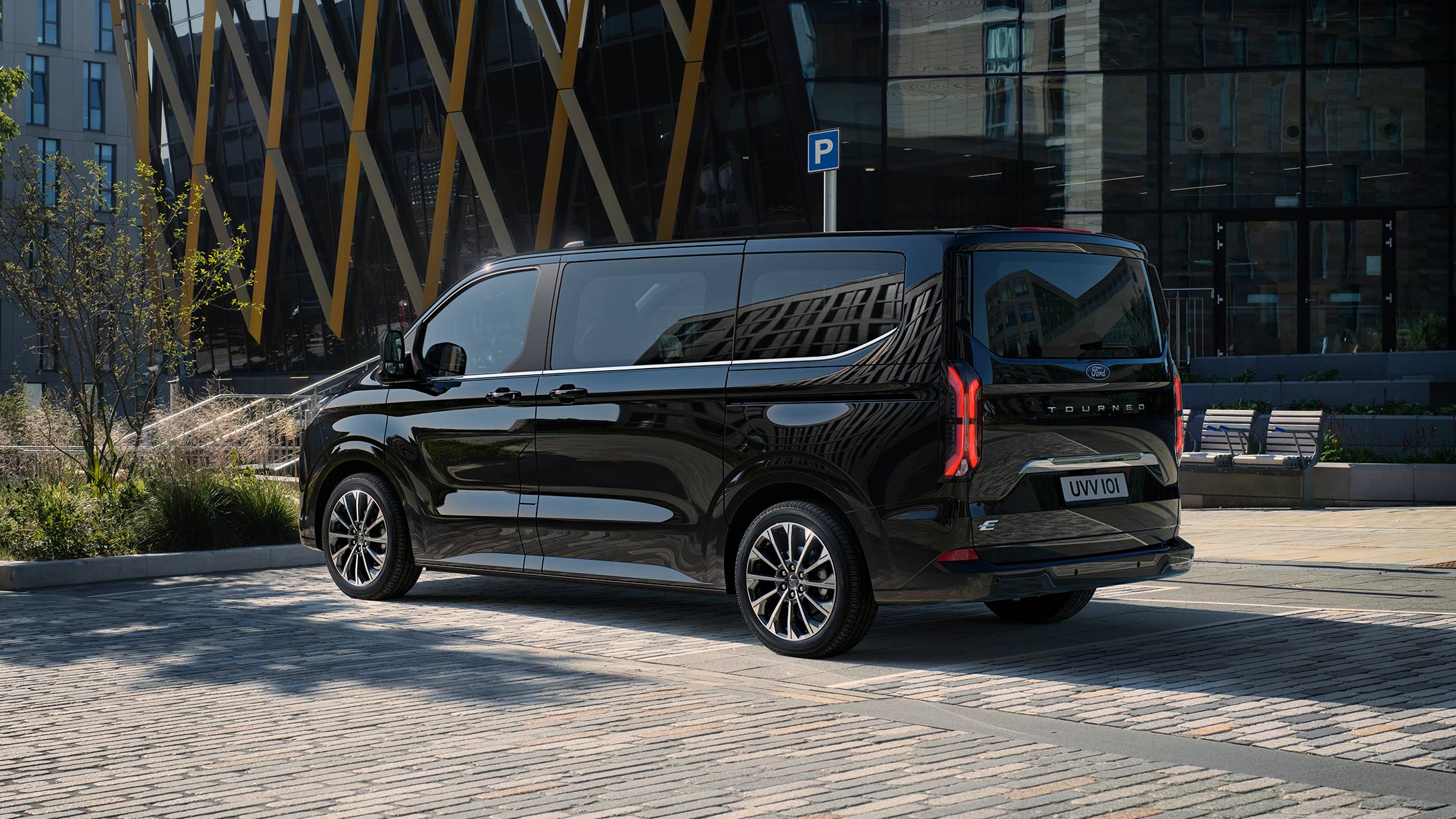 Ford Tourneo Custom in Schwarz in  der ¾-Heckansicht geparkt auf einem Parkplatz vor modernen Gebäuden