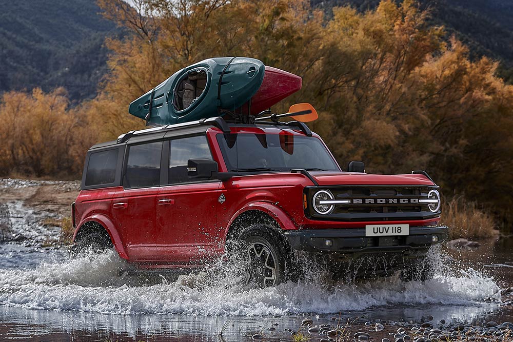Seitenansicht eines roten Ford Bronco, der durch flaches Wasser in einer Berglandschaft fährt.