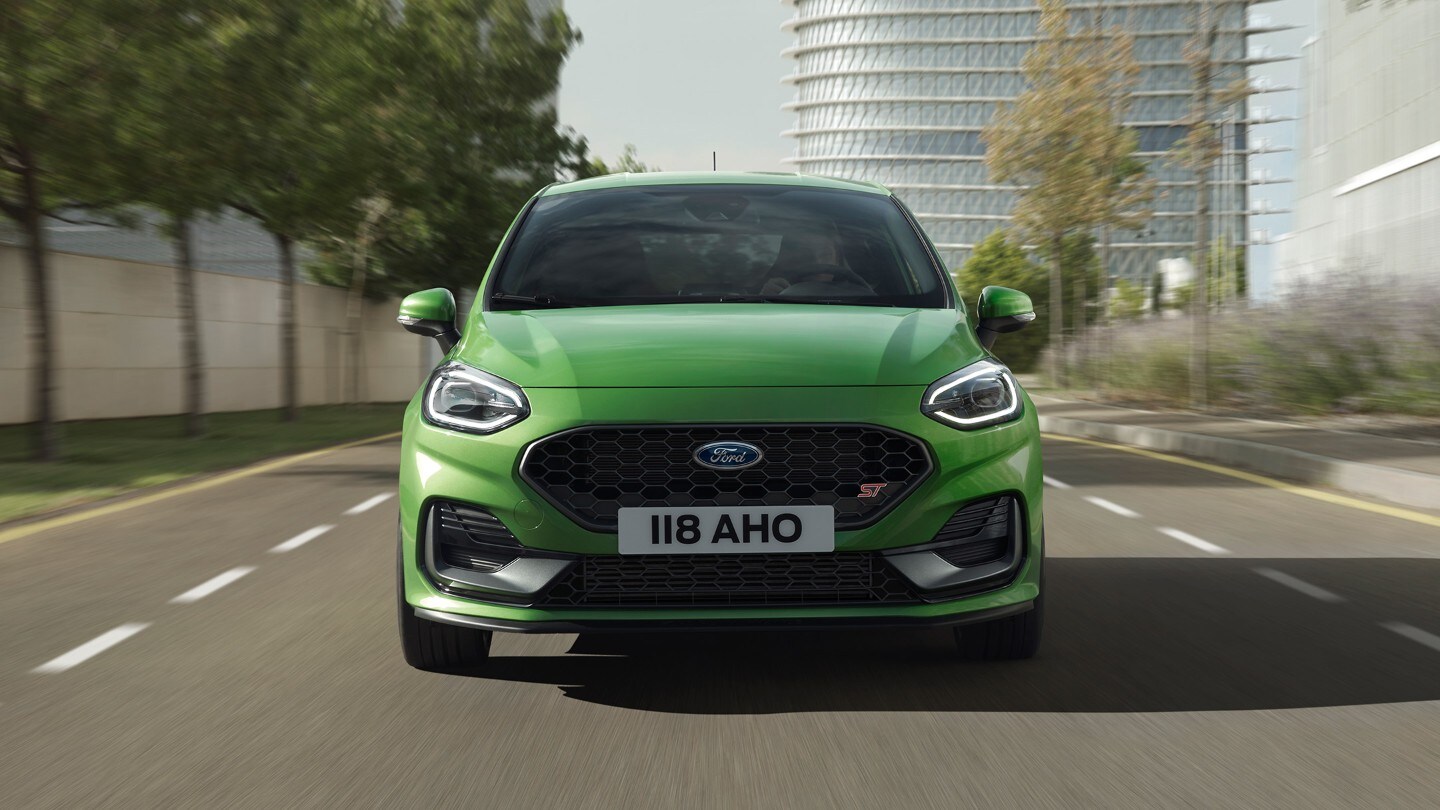 Ford Fiesta ST in Grün. Frontansicht auf Strasse vor Bäumen und modernem Gebäude.