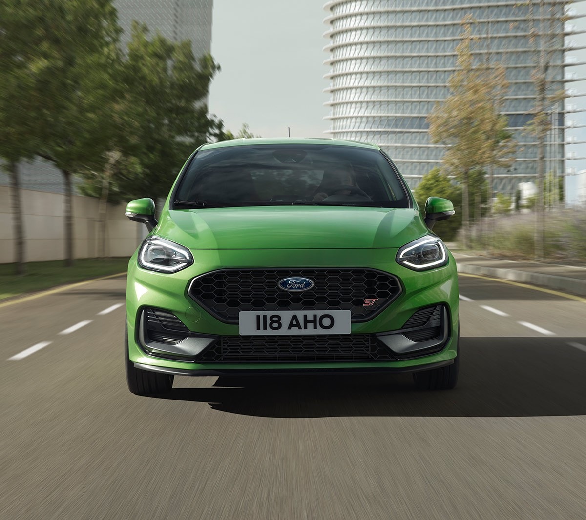 Ford Fiesta ST in Grün. Frontansicht, auf Strasse vor Bäumen und modernem Gebäude fahrend.