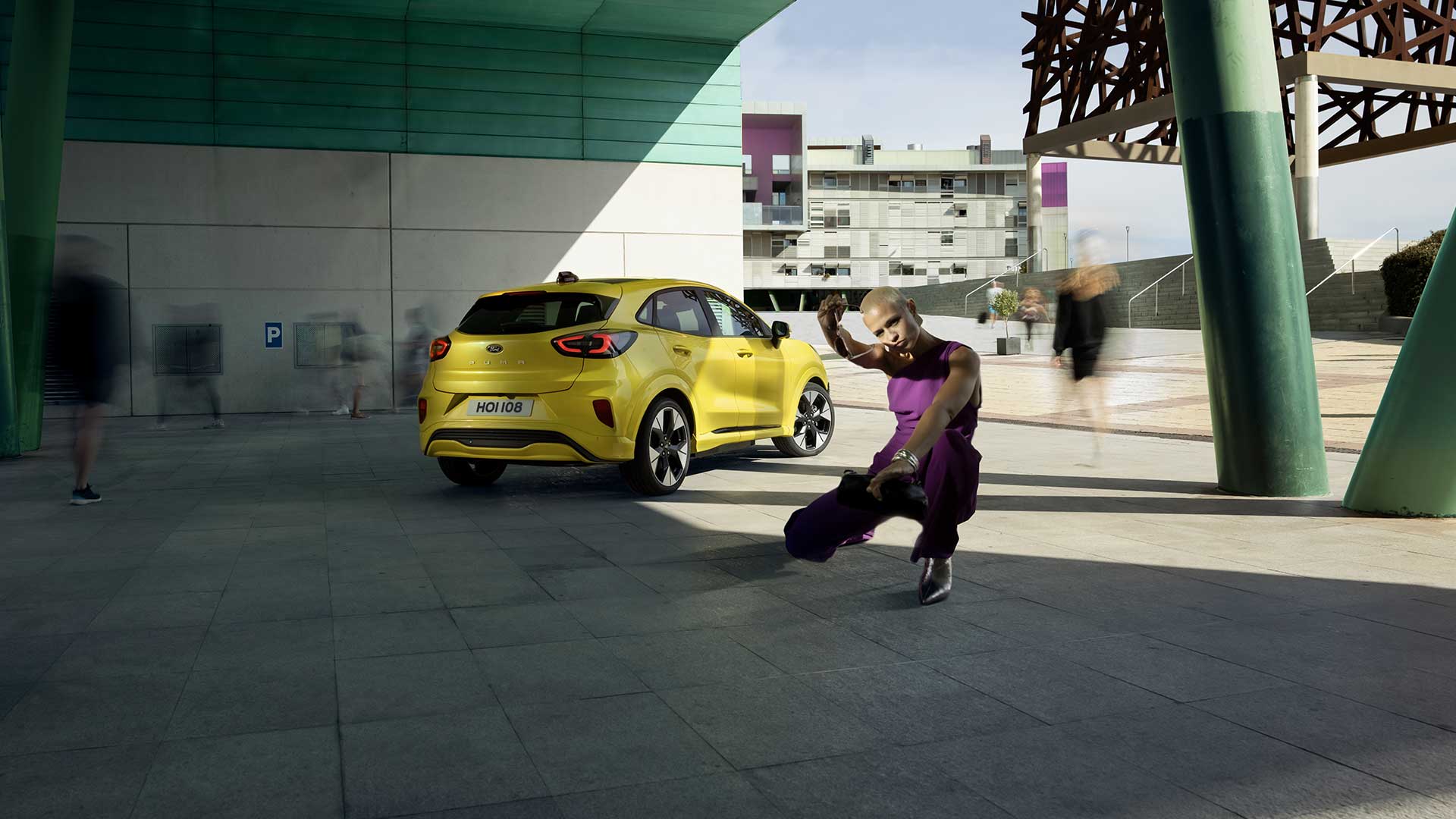 Heckansicht eines vollelektrischen Ford Puma Gen-E® in Gelb, der unter einer Brücke parkiert ist, neben einer violett gekleideten Person.