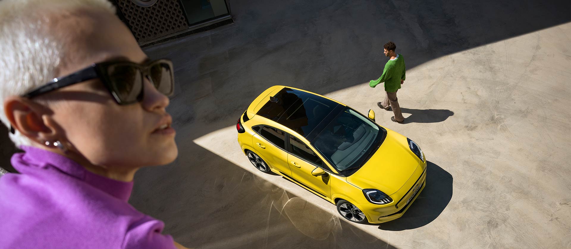 Vollelektrischer Ford Puma Gen-E® in Gelb aus der Vogelperspektive. Im Vordergrund ist eine violett gekleidete Person zu sehen. 