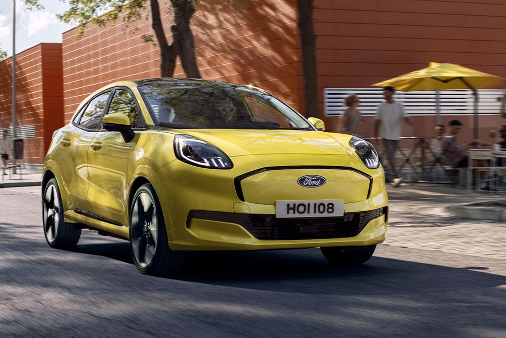 Frontansicht eines vollelektrischen Ford Puma Gen-E® in Gelb, der auf einer Stadtstrasse fährt.