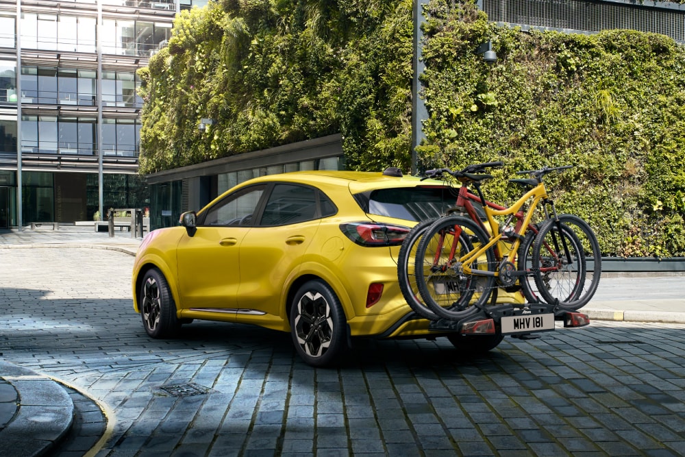 Ein gelber Ford Puma Gen-E, geparkt in einem urbanen Umfeld, mit zwei an einem Heckfahrradträger montierten Fahrrädern.