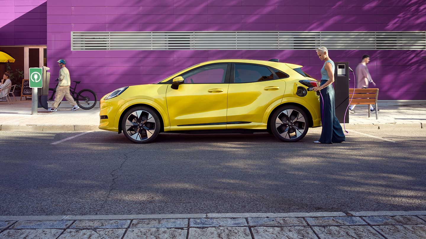 Seitenansicht eines parkierten vollelektrischen Ford Puma Gen-E® in Gelb, der an eine öffentliche Ladestation angeschlossen ist.