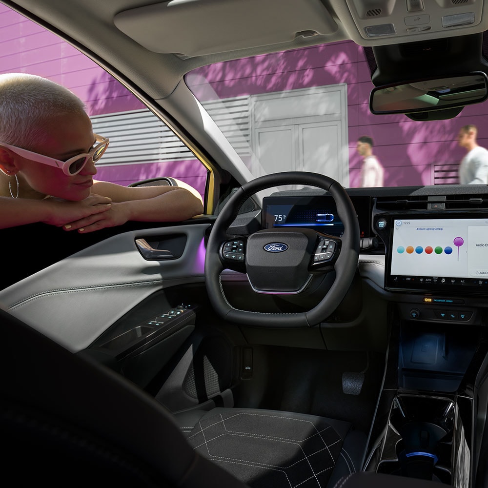 Innenraum des vollelektrischen Ford Puma Gen-E®, während eine Person den Infotainment-Bildschirm durch das offene Fenster der Fahrertür betrachtet.