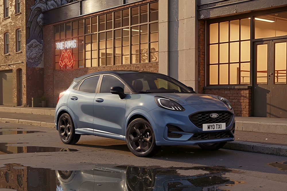 Ein blauer Ford Puma parkt bei Sonnenuntergang auf einer städtischen Strasse mit Pfützen, die die Gebäude widerspiegeln.