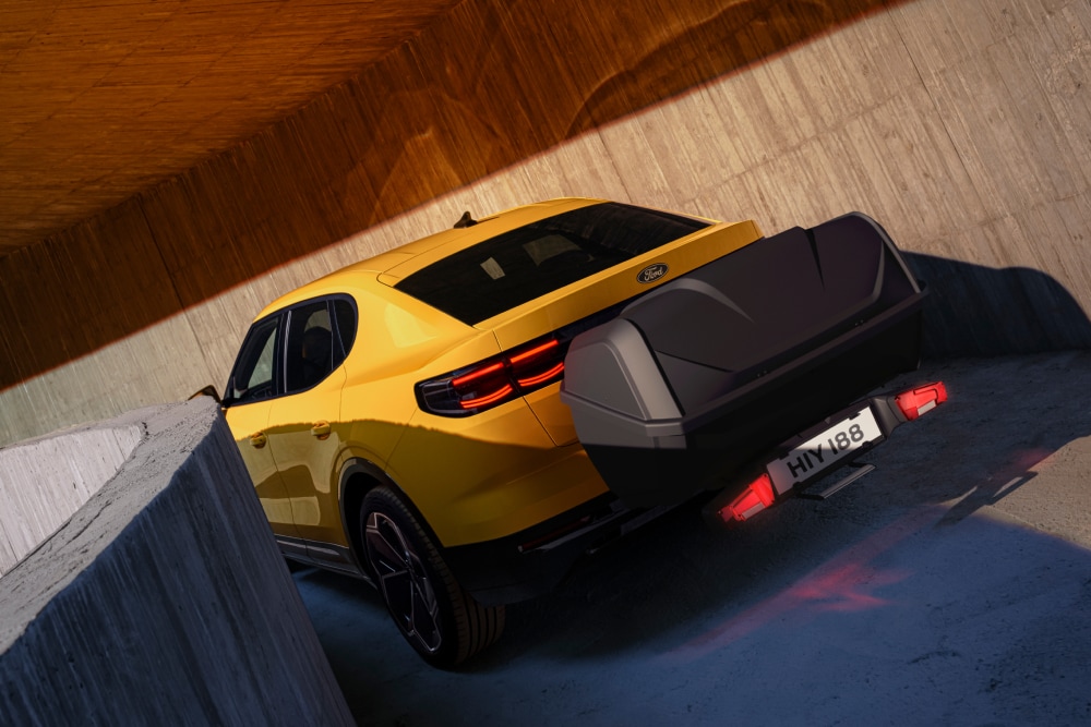 Rückansicht eines gelben Ford Capri, geparkt in einem modernen Betonbau, mit Gepäckbox, die mit einem Gepäckbox-Träger am Heck befestigt ist.